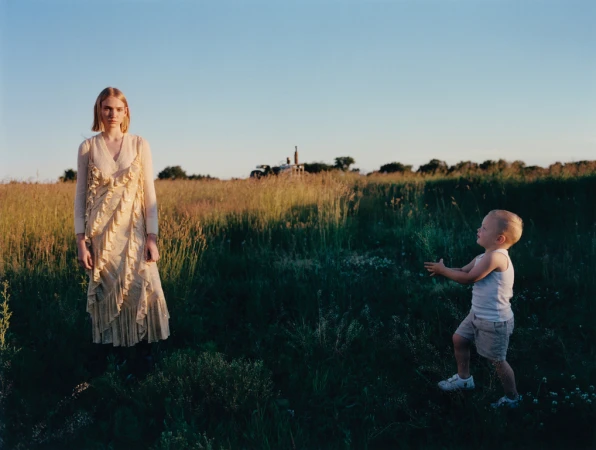 A woman in a long cream ruffled dress stands in a grassy field with a toddler reaching toward her under a clear sky.