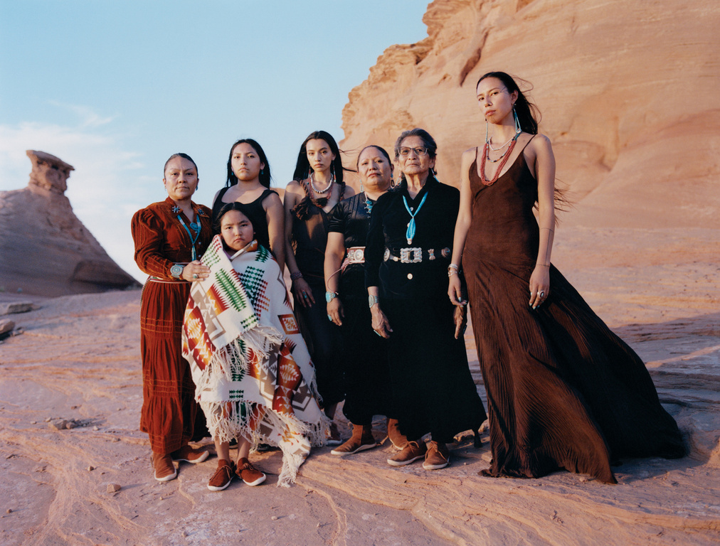 Seven Indigenous women and a child stand together in a desert landscape with red rock formations, wearing traditional clothing and jewelry.