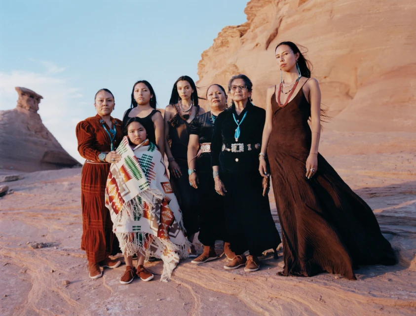 Seven Indigenous women and a child stand together in a desert landscape with red rock formations, wearing traditional clothing and jewelry.