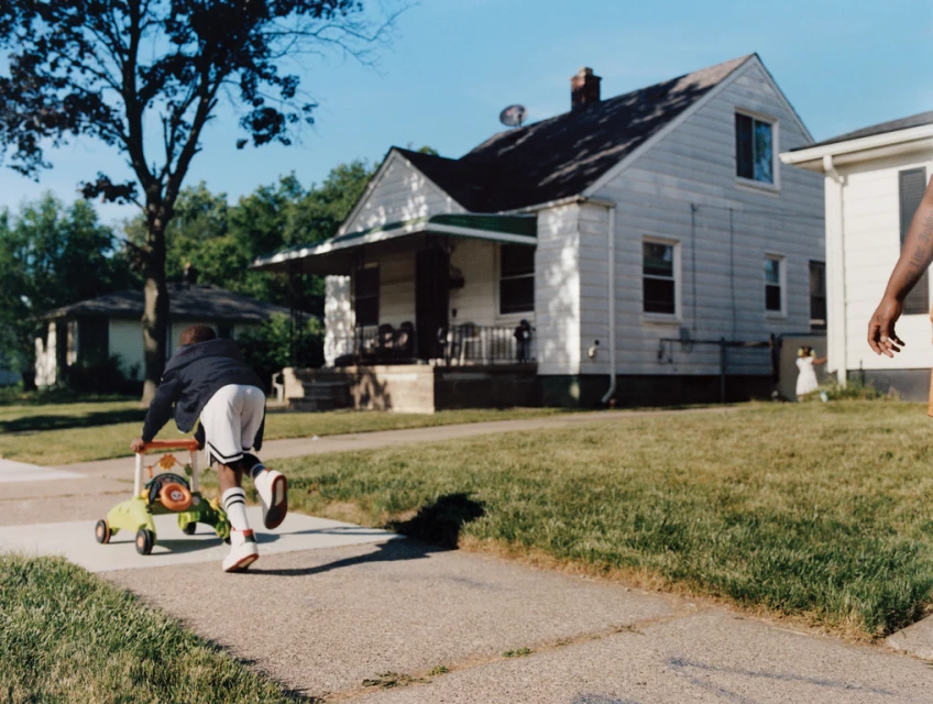 Young child pushes a baby walker down a sidewalk on a sunny day in a residential area with houses and trees in the background.