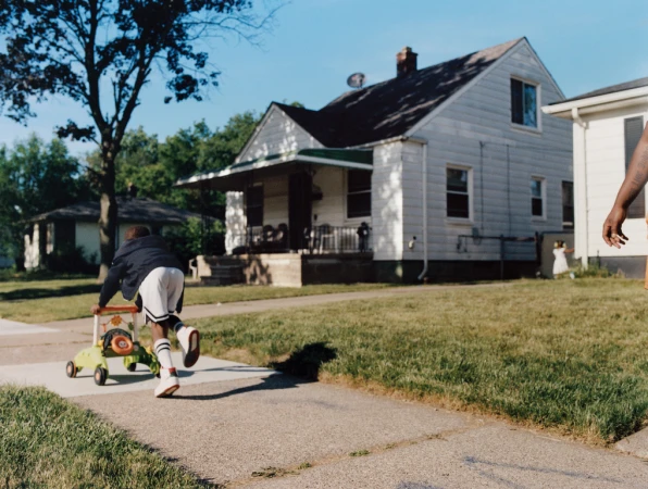 Young child pushes a baby walker down a sidewalk on a sunny day in a residential area with houses and trees in the background.