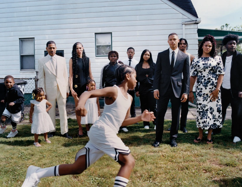 A large group of African American people in formal attire stand on a lawn outside of a house, with a child running toward the camera.