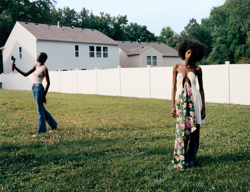 Two Black women pose outdoors on a grassy lawn with white houses in the background; one holds a camera, the other wears a unique dress.