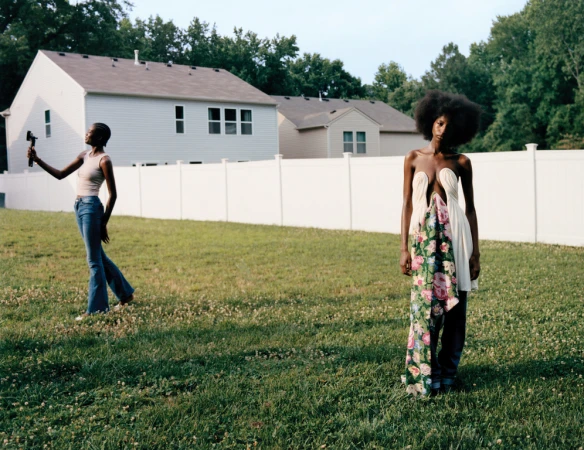 Two Black women pose outdoors on a grassy lawn with white houses in the background; one holds a camera, the other wears a unique dress.