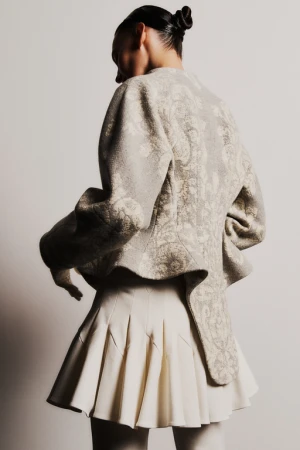 Model wearing a patterned structured jacket and pleated skirt against a plain studio background
