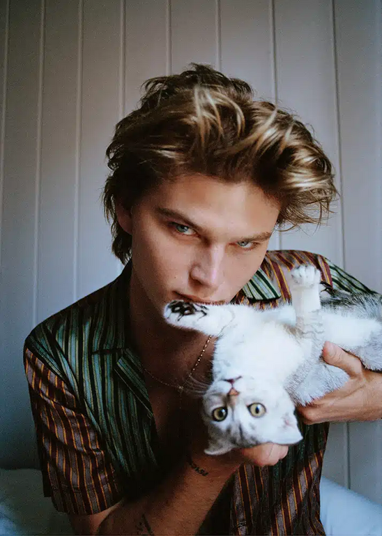 An adult male with blond hair and blue eyes is holding a gray tabby cat in a portrait photo taken indoors against a wall.