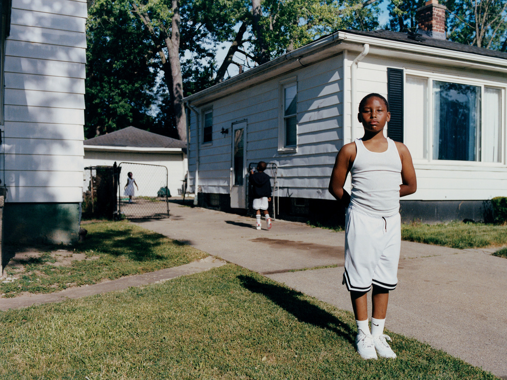 Portrait of a young Black boy in a white tank top and basketball shorts standing in front of a suburban home with a grassy lawn.
