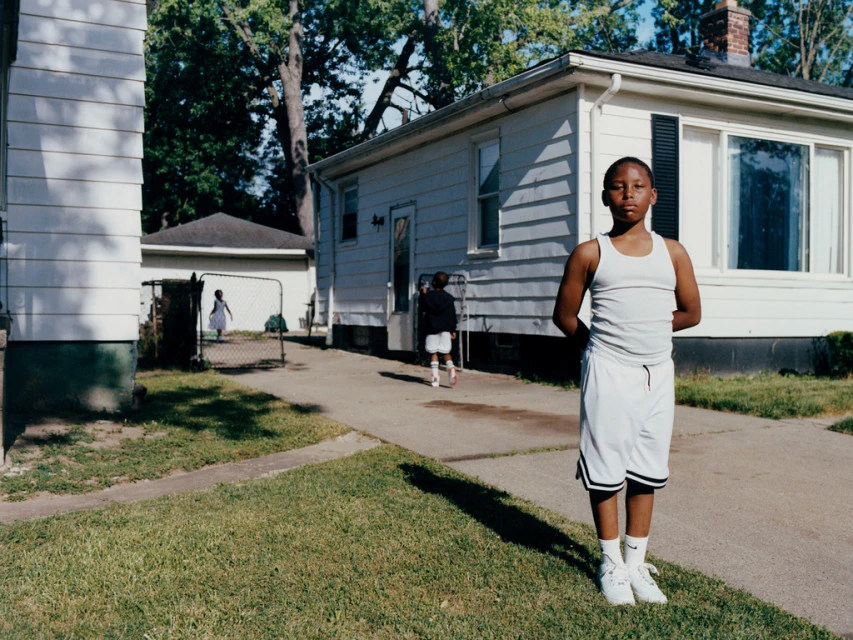 Portrait of a young Black boy in a white tank top and basketball shorts standing in front of a suburban home with a grassy lawn.