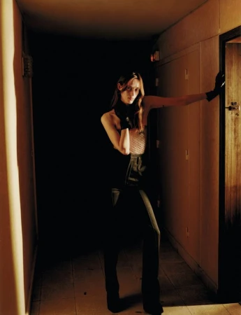 Woman with long brown hair wearing a mesh top and dark gloves posing in a narrow lit hallway by a doorway.