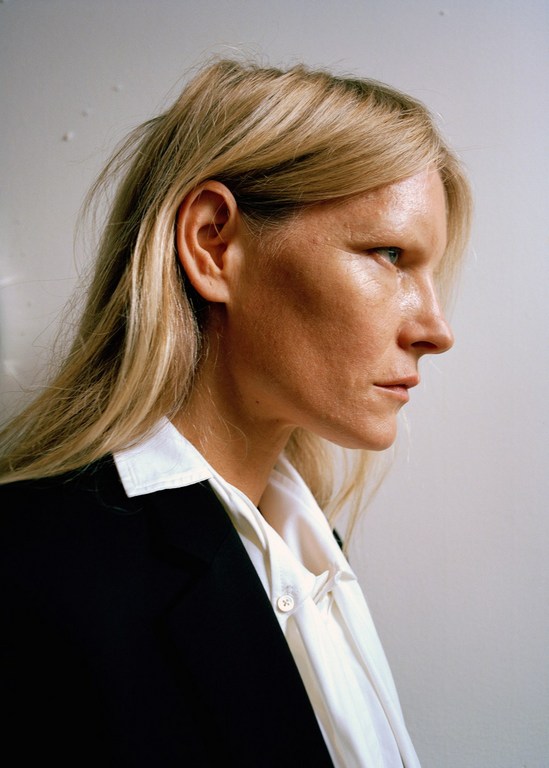 Profile portrait of a blonde adult woman in a black blazer and white button-down shirt against a white wall, showcasing beauty and fashion.