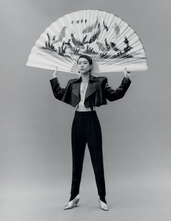 Woman in black jacket and pants poses with an open fan in front of a neutral background in a studio, fashion shoot.