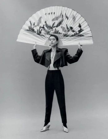 Woman in black jacket and pants poses with an open fan in front of a neutral background in a studio, fashion shoot.