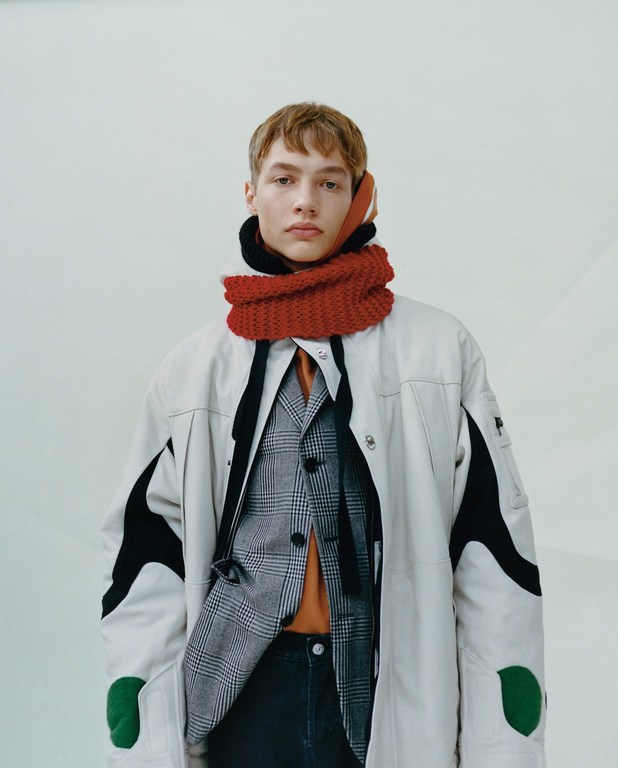 Fashion portrait of a young adult male with fair skin wearing a tan jacket and a red knit scarf indoors, medium shot, eye-level angle.