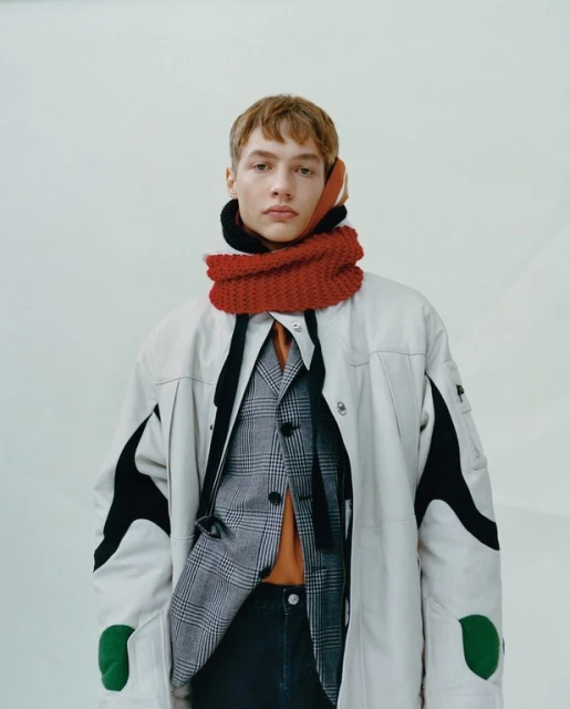 Fashion portrait of a young adult male with fair skin wearing a tan jacket and a red knit scarf indoors, medium shot, eye-level angle.