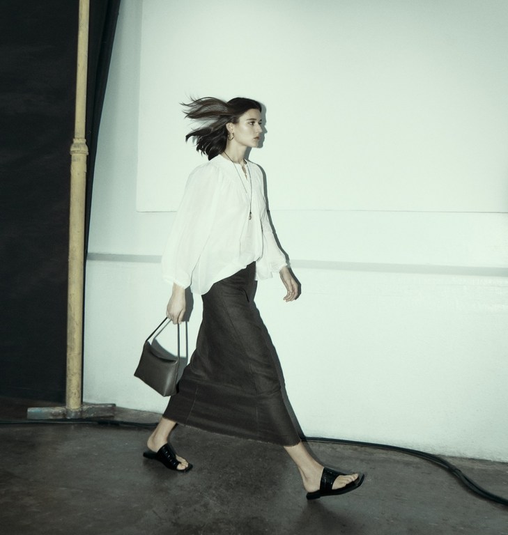 Woman walking past a pale wall wearing a white long-sleeve blouse and long dark skirt holding a handbag