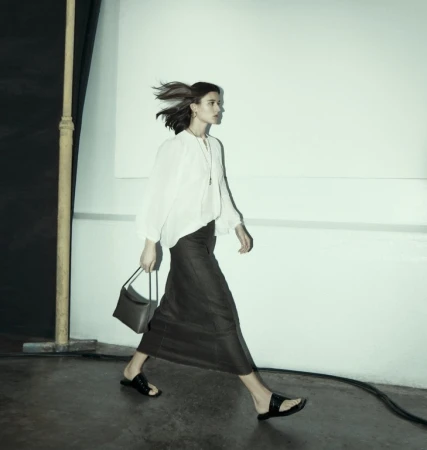 Woman walking past a pale wall wearing a white long-sleeve blouse and long dark skirt holding a handbag