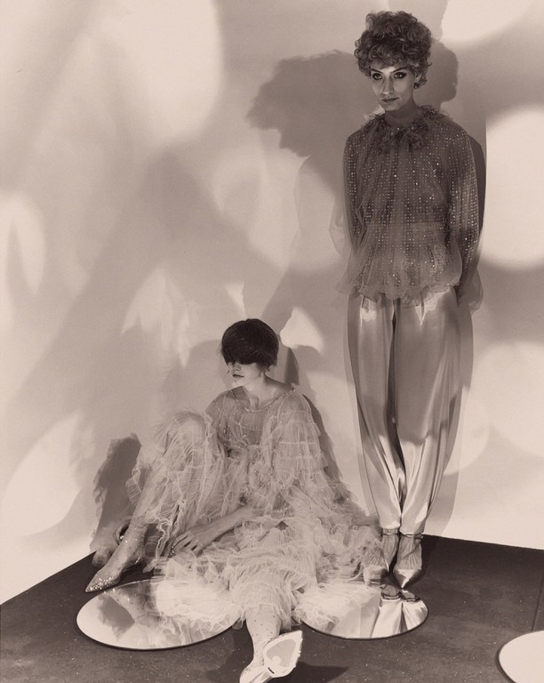 Two women in frilly, layered tops and bottoms, one standing and one sitting on mirror platforms against a mottled wall in sepia tones.