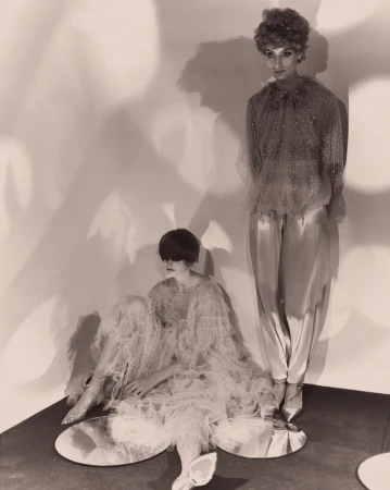 Two women in frilly, layered tops and bottoms, one standing and one sitting on mirror platforms against a mottled wall in sepia tones.
