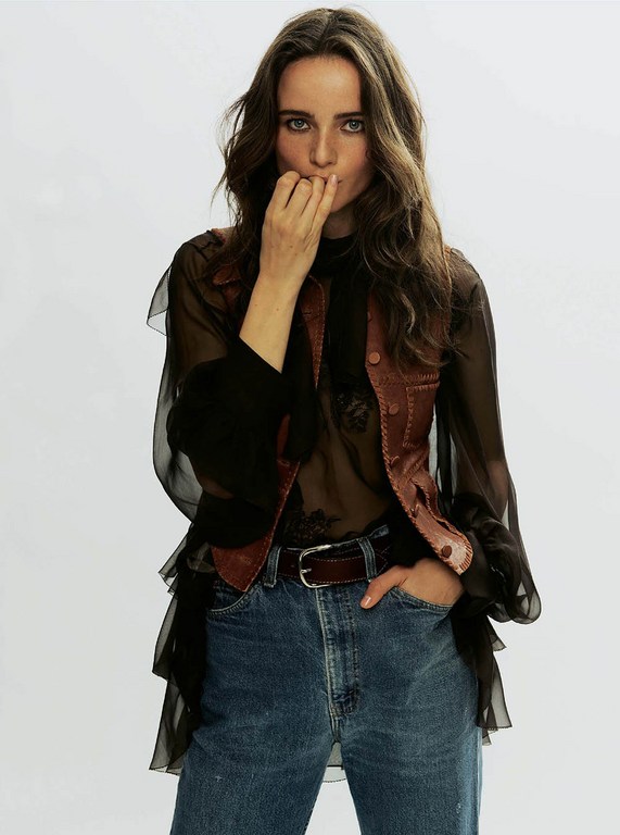 A brunette woman poses in a sheer top, vest, and jeans against a white backdrop, with hand near mouth, looking directly at the camera.