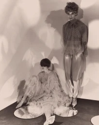 Two models in sheer ruffled blouses and shiny trousers posing near circular mirrors in a studio lit with dappled patterns.