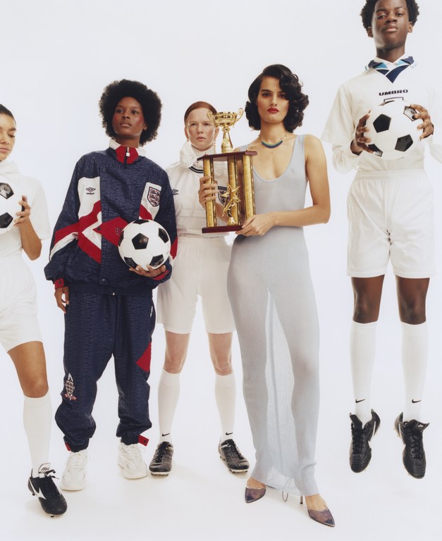 Group of five diverse people in athletic wear posing in a photo studio with soccer balls and a trophy in a light and airy setting.