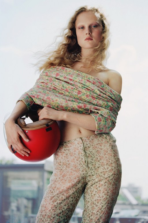 Blonde woman in floral pants and top holding a red helmet against a city background with neutral expression, soft lighting.