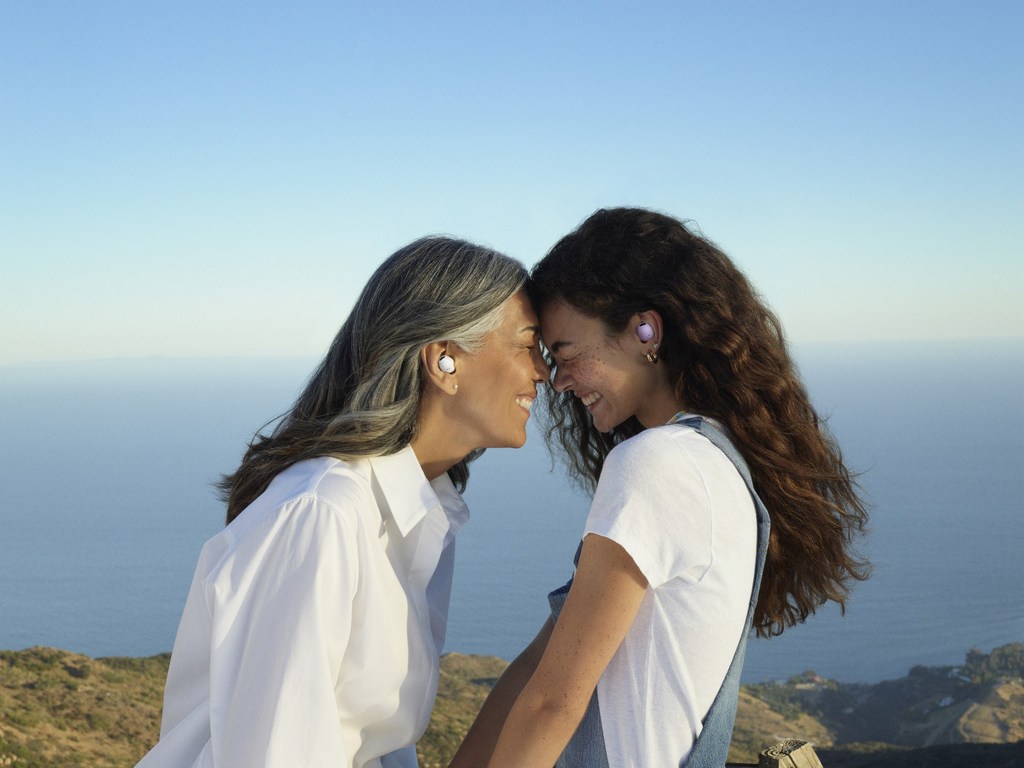 A smiling adult woman and a young woman with foreheads touching outdoors with a sea and sky background wearing wireless earbuds.