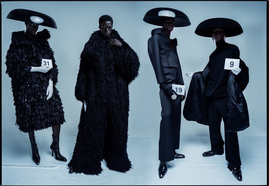 Four fashion models showcase avant-garde black couture, featuring oversized hats and dramatic textures, against a light background in studio.