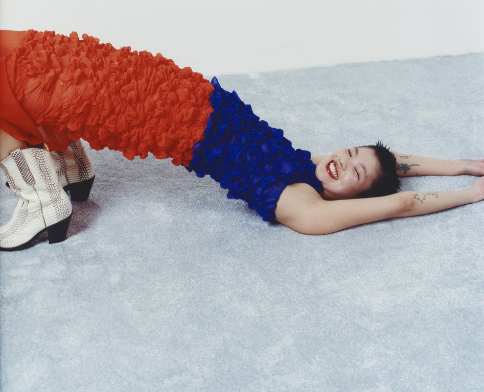 Smiling woman in blue and orange textured dress arches her back on a blue surface, showcasing a playful pose with white snakeskin boots.