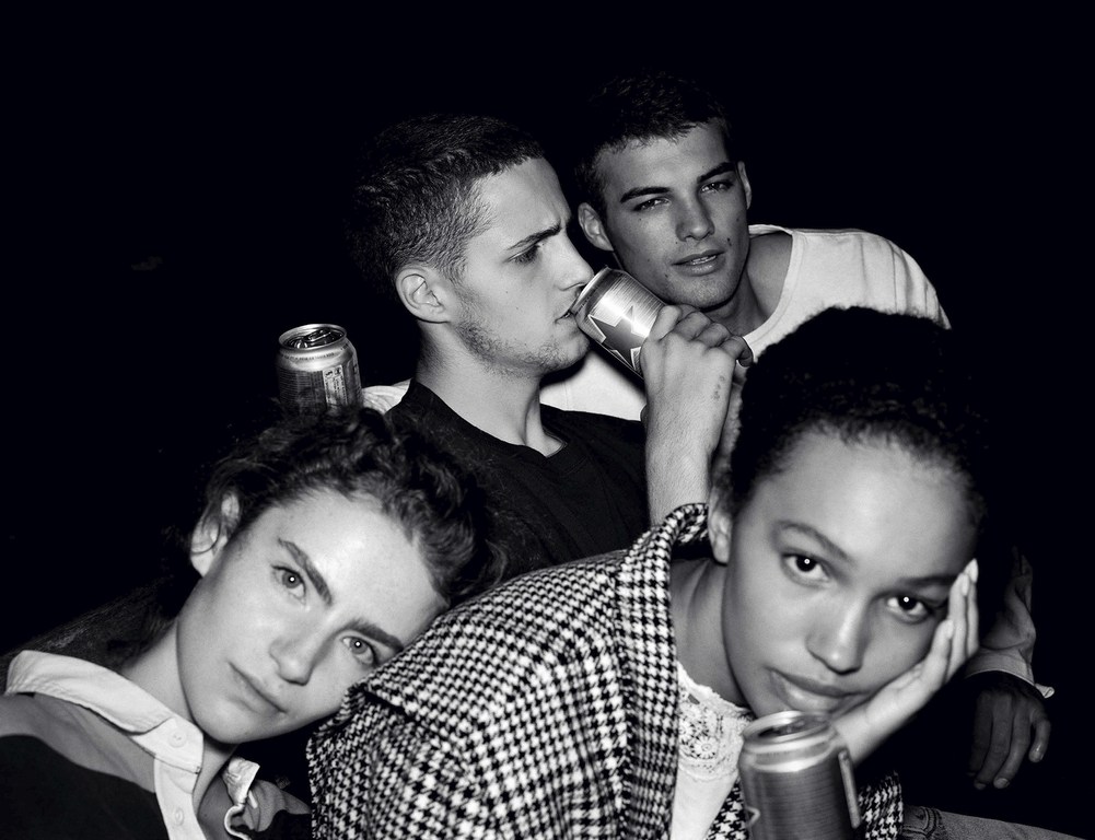 Four young adults pose indoors, holding aluminum cans in a black and white image with two women and two men in casual clothing.