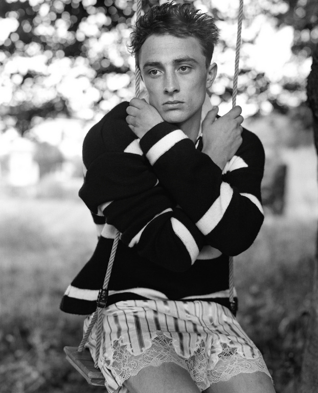 Monochrome portrait of a young adult man sitting on a swing in a rural setting wearing a striped shirt and lace-trimmed shorts.