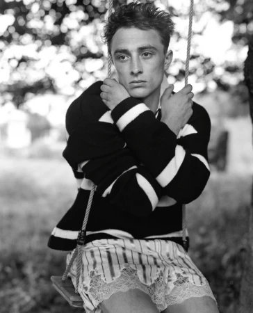 Monochrome portrait of a young adult man sitting on a swing in a rural setting wearing a striped shirt and lace-trimmed shorts.