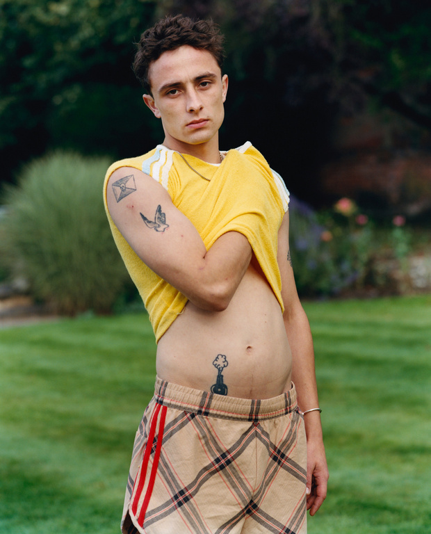 Young adult male poses outdoors with tattoos visible on arm, chest and abdomen, short shorts and yellow t-shirt, medium shot.