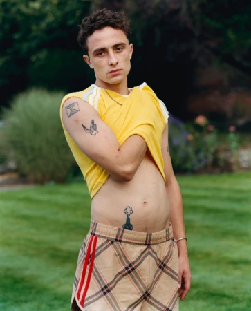 Young adult male poses outdoors with tattoos visible on arm, chest and abdomen, short shorts and yellow t-shirt, medium shot.