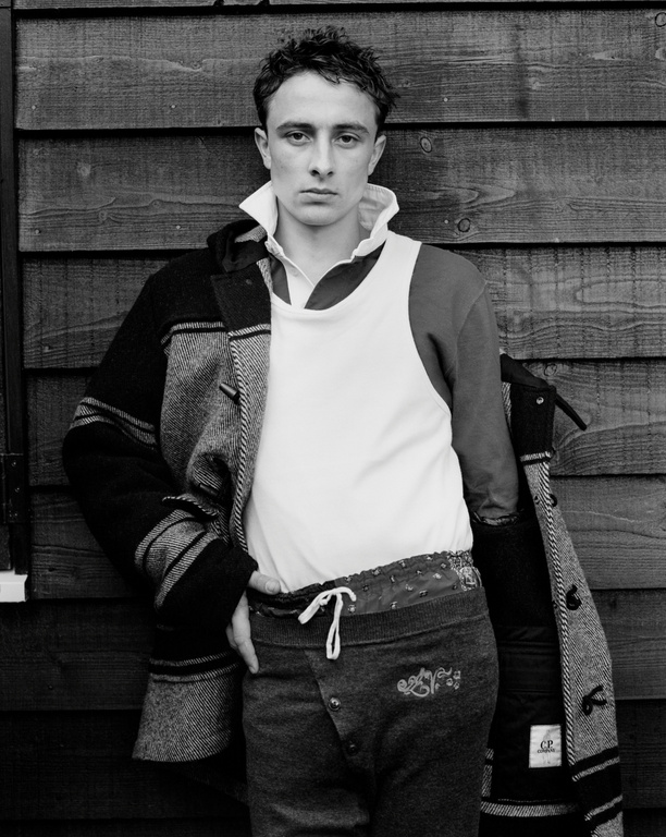 Monochrome portrait of a young man posing in jacket, white shirt, and patterned pants, standing against a wooden wall outdoors.