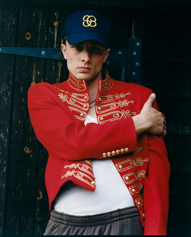Young adult male in a red hussar jacket and blue baseball cap poses indoors against a dark wooden background with metal hardware.
