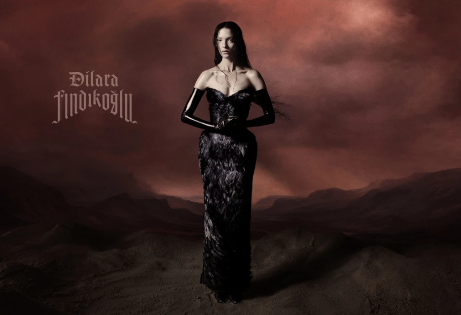 A woman in a dark gown stands in a surreal landscape against a red sky, a striking fashion portrait with a dramatic, gothic aesthetic.