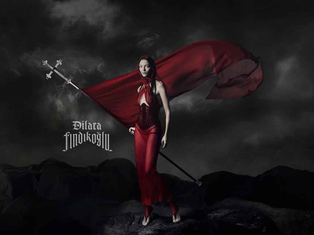Woman in a flowing red dress and hood holds a decorative flagstaff in a dramatic, dark landscape with cloudy skies above.