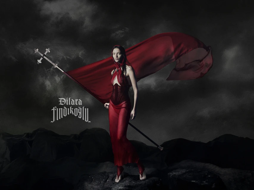 Woman in a flowing red dress and hood holds a decorative flagstaff in a dramatic, dark landscape with cloudy skies above.