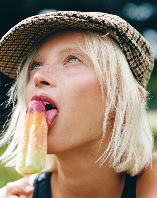 Close-up portrait of a blonde woman with a houndstooth cap, her tongue extended to an orange, pink, and white popsicle in an outdoor setting.