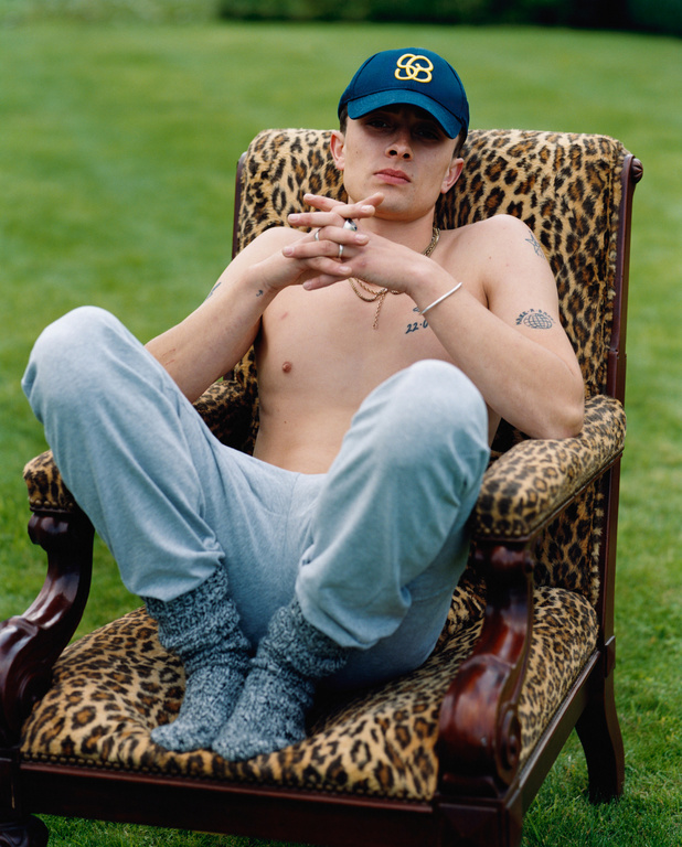 Young adult male sits in an armchair with a leopard print in a grassy outdoor setting, wearing a baseball cap and grey sweatpants.