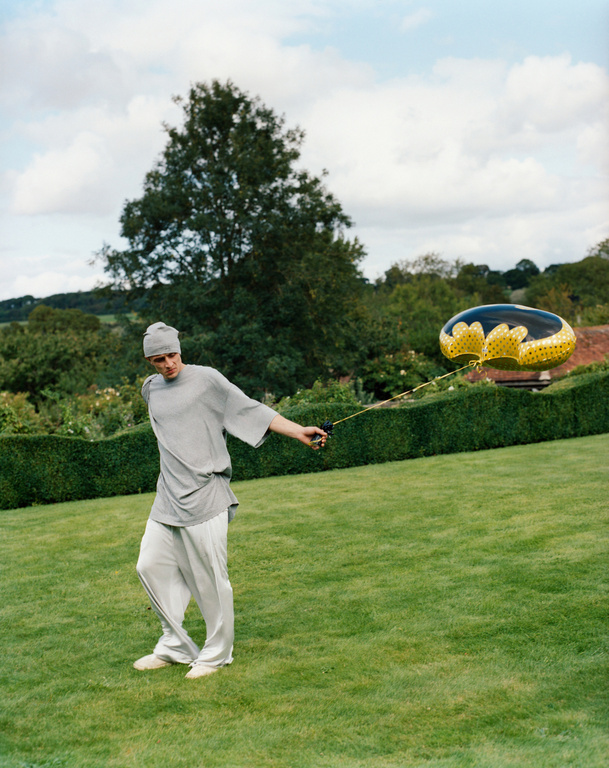 Man in grey tracksuit pulls a balloon on a green lawn, with trees, cloudy sky and topiary in the background on a summer day.