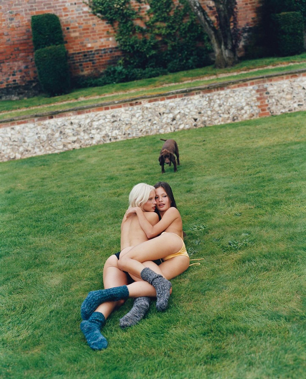 Two women embrace on green lawn wearing knee socks, bathed in natural light, with a dog in the background and brick building.