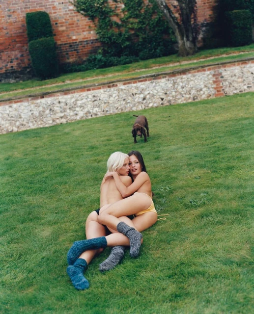 Two women embrace on green lawn wearing knee socks, bathed in natural light, with a dog in the background and brick building.