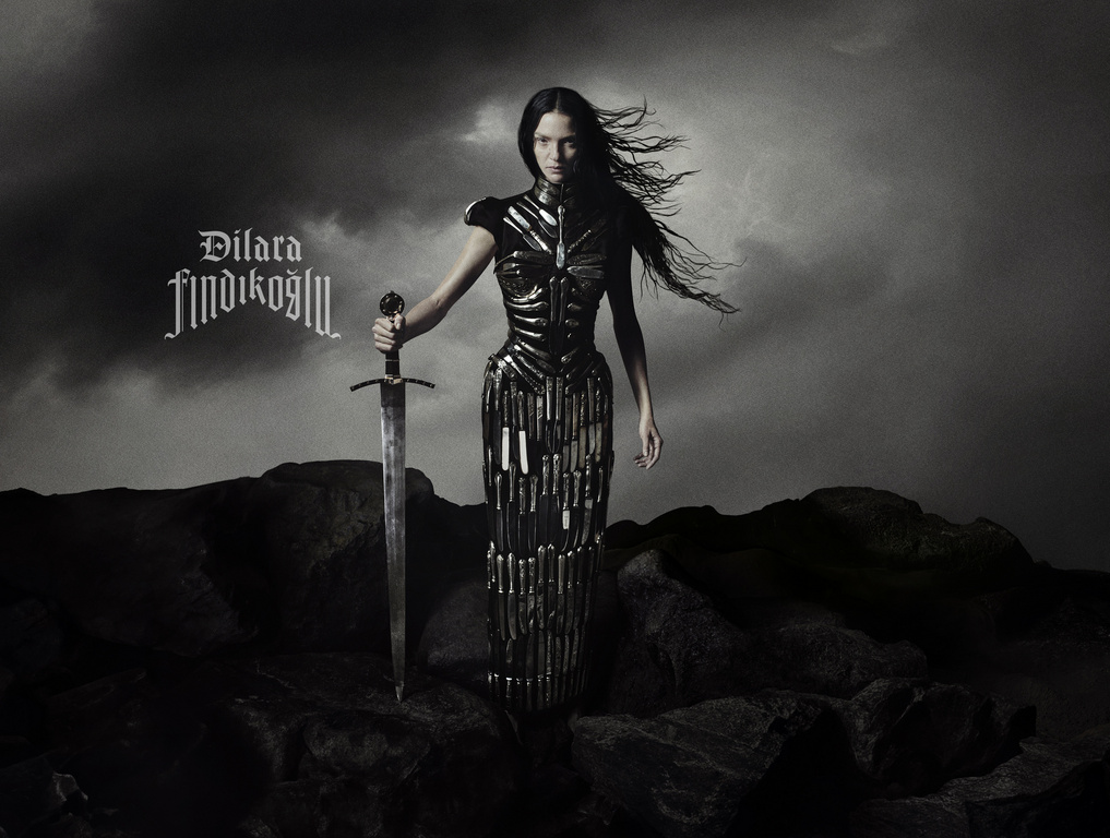 A woman in an elaborate dark dress stands in an outdoor landscape with a sword in hand against a cloudy, monochrome backdrop.