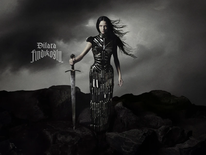 A woman in an elaborate dark dress stands in an outdoor landscape with a sword in hand against a cloudy, monochrome backdrop.