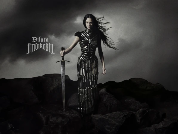 A woman in an elaborate dark dress stands in an outdoor landscape with a sword in hand against a cloudy, monochrome backdrop.