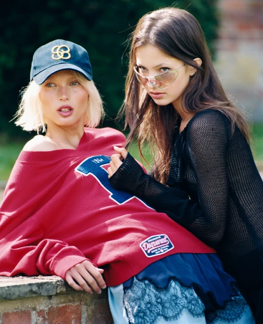 Two female models in an outdoor setting. One with a blonde bob wears a baseball cap and red sweatshirt. The other wears sunglasses.