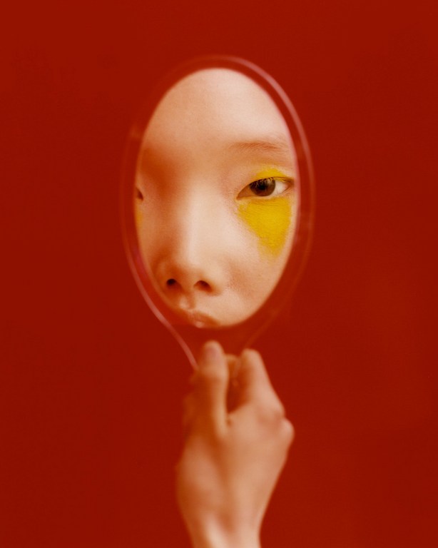 Close-up portrait reflected in hand mirror against red background, featuring woman with yellow eyeshadow in a studio setting.