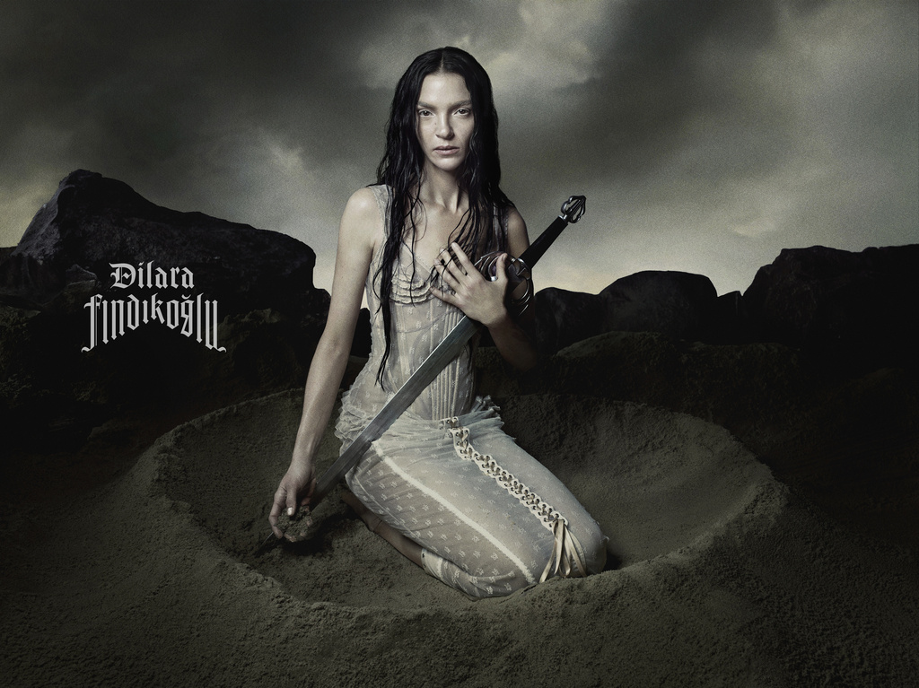 Dramatic portrait of a woman kneeling in sand, holding a sword, under a stormy sky. Dark, moody aesthetic with a gothic-inspired vibe.